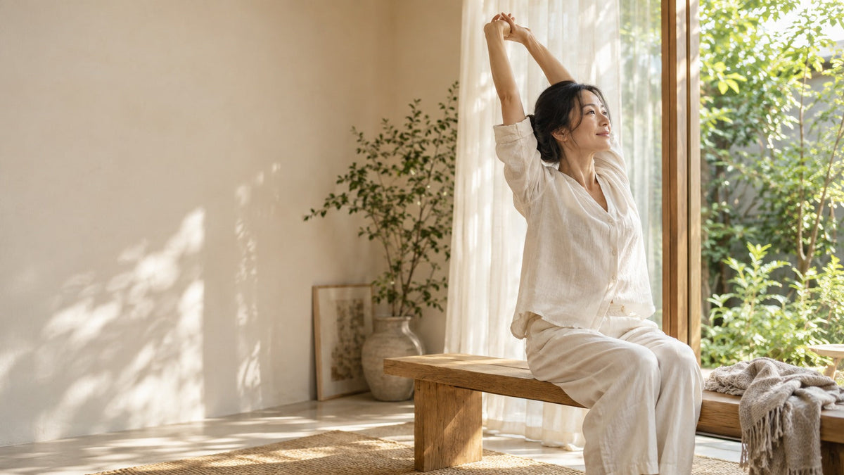 Woman stretching her arms overhead while seated on a wooden bench beside a sunlit window.